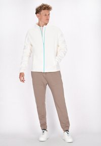 Witte zip-up jas met turkoois rits, gecombineerd met beige sweatpants. Het model draagt witte sneakers en toont een ontspannen pasvorm en casual stijl.