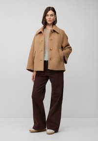 Kobieta ubrana w płaszcz w kolorze camel, beżowy sweter, ciemne szerokie spodnie i beżowe sneakersy, stojąca na tle jednolitego szarego tła.