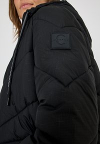 Sort vinterjakke med et chevron-ternet mønster. Har et tekstureret badge på ærmet og en hætte med snore.