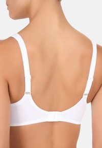 Schiena nuda di una donna che indossa un reggiseno bianco regolabile con chiusura a gancio su uno sfondo chiaro e semplice.
