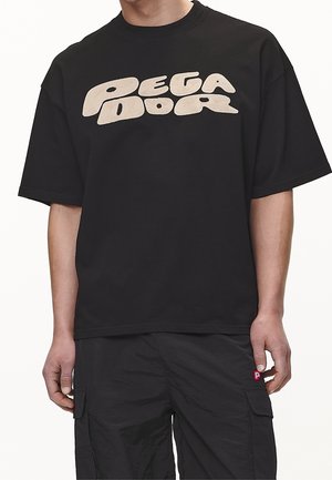 Person trägt ein schwarzes, übergroßes T-Shirt mit beigem, stilisiertem „PEGADOR“-Schriftzug über der Brust und schwarze Cargohosen mit Seitentaschen.