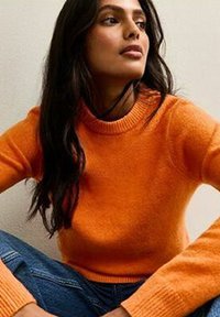 Femme aux longs cheveux noirs portant un pull orange et un jean bleu, assise avec les bras reposant sur les genoux, regardant légèrement vers le haut.