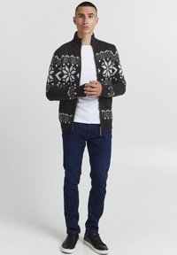 Schwarze Strickjacke mit weißem Schneeflockenmuster, hohem Kragen, Reißverschluss vorne, getragen über einem weißen T-Shirt; kombiniert mit blauen Jeans und schwarzen Sneakers.