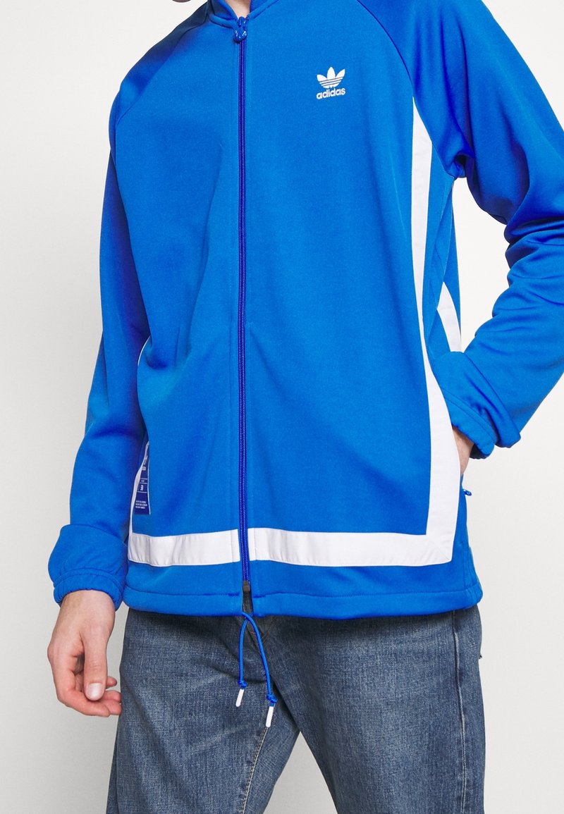 Sininen huppari valkoisilla yksityiskohdilla, vetoketjusuljenta, pehmeä kangas, resorikahleet ja kiristysnaru helmassa. Adidas-logo rinnassa.