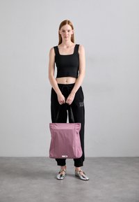 Kvinne med rødt hår i svart crop top og joggebukser som holder en stor rosa tote bag, står på et grått gulv mot en ensfarget vegg.