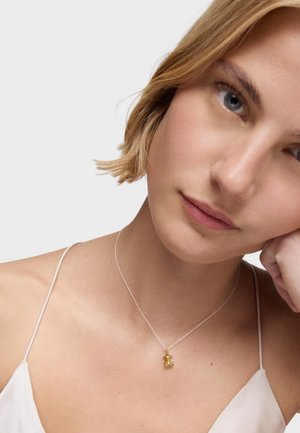 Jeune femme aux cheveux blonds courts portant un haut à fines bretelles blanc et un délicat collier en argent avec un petit pendentif ours en or.