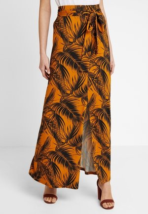 Longue jupe orange avec motif feuilles tropicales noires, fente avant, ceinture nouée à la taille, portée avec des sandales marron à talons ouverts.