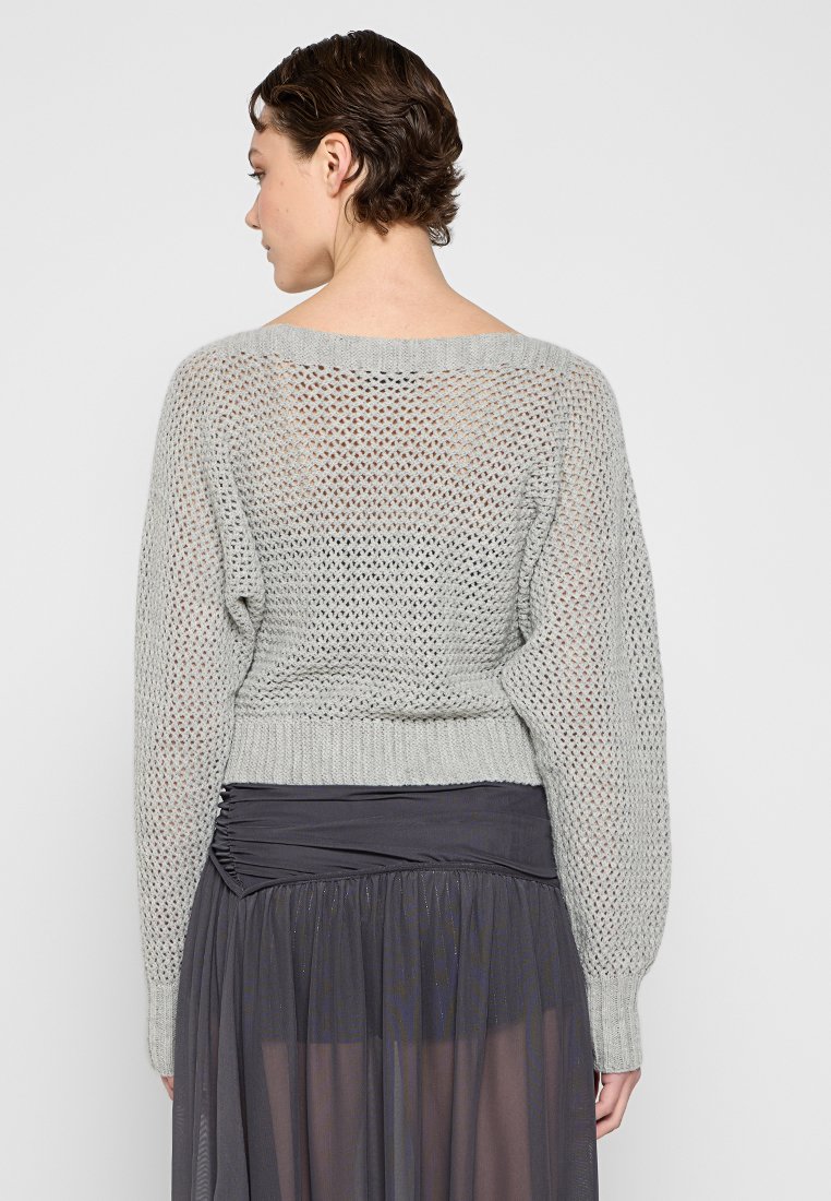 Hellgrauer, gestrickter Pullover mit lockerem, offenem Gewebe und langen Ärmeln, die bis zur Taille reichen. Getragen über einem dunklen, mehrlagigen Rock.