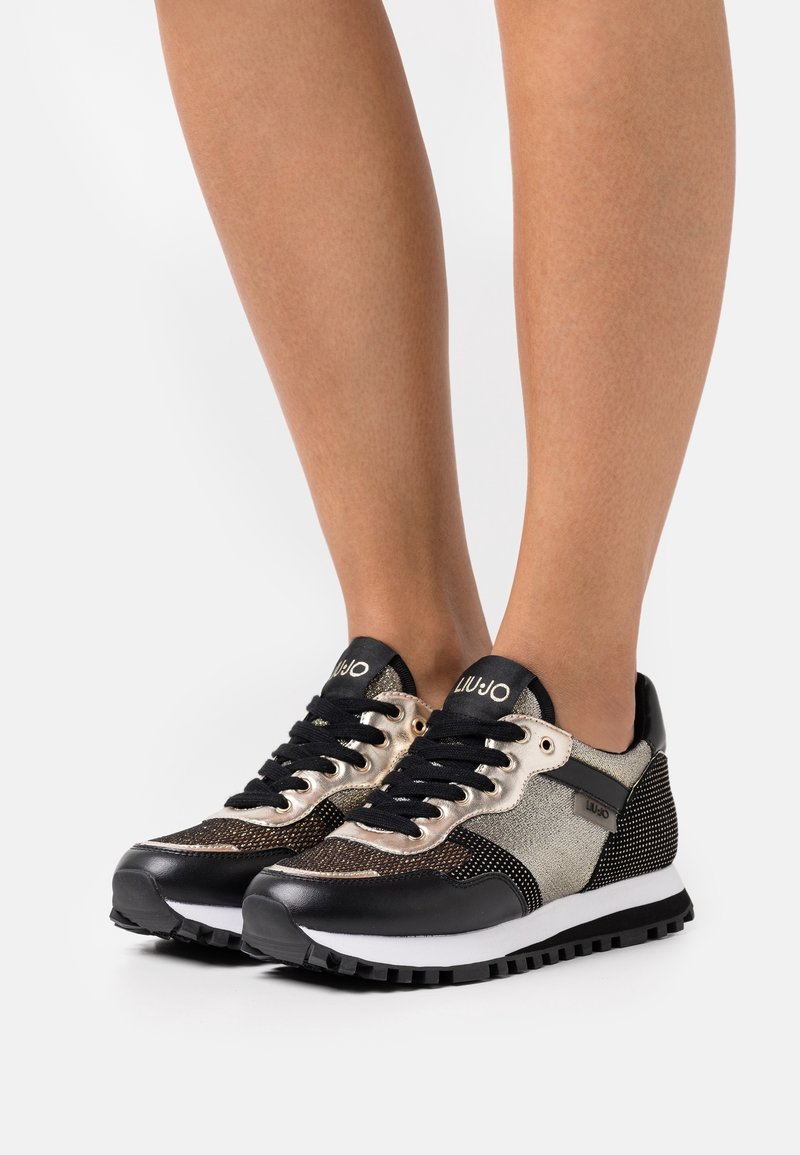 Paire de baskets Liu Jo noires, dorées et beiges avec des lacets noirs portées sur des jambes nues sur fond blanc uni.