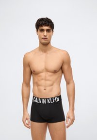 Barbarklädd man med kort lockigt hår, iförd svarta Calvin Klein kalsonger, som står framför en enkel ljus bakgrund.