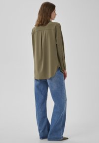 Eine Frau mit schulterlangen braunen Haaren trägt ein olivgrünes Langarmshirt und weite blaue Jeans und steht vor einem einfarbigen hellen Hintergrund.