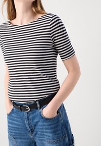 Camiseta de rayas en blanco y negro con cuello redondo y mangas cortas, combinada con pantalones vaqueros de mezclilla azul de talle alto y un cinturón negro.