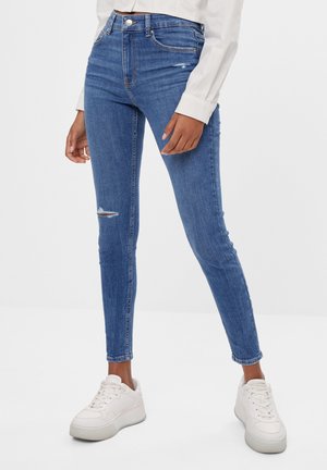 Niebieskie jeansy w fasonie slim-fit z lekkim przetarciem na lewym kolanie, połączone z białymi sneakersami na platformie i białą koszulą z długim rękawem.