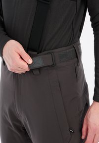 Dunkelgraue Hose mit verstellbaren Trägern, ausgestattet mit einem Klettverschluss und mehreren Taschen. Glatte Textur und taillierte Passform.