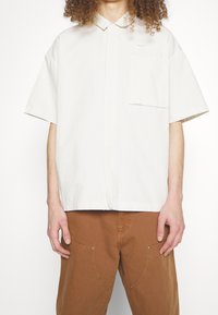 Krátký rukáv, bílá košile na knoflíky vyrobená z bavlny, s jednou náprsní kapsou a jemně vyšitým logem značky na levé straně.