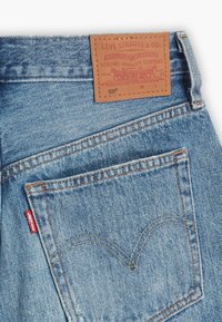 Ljust blå denimjeans med en läderlapp med röd och guldig text, en liten röd etikett och ikonisk bakfickssöm.