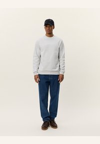 Sudadera gris con cuello redondo, de tela ligera y puños de punto, combinada con unos vaqueros de denim azul holgados y zapatos marrón oscuro. Gorra negra con logo.