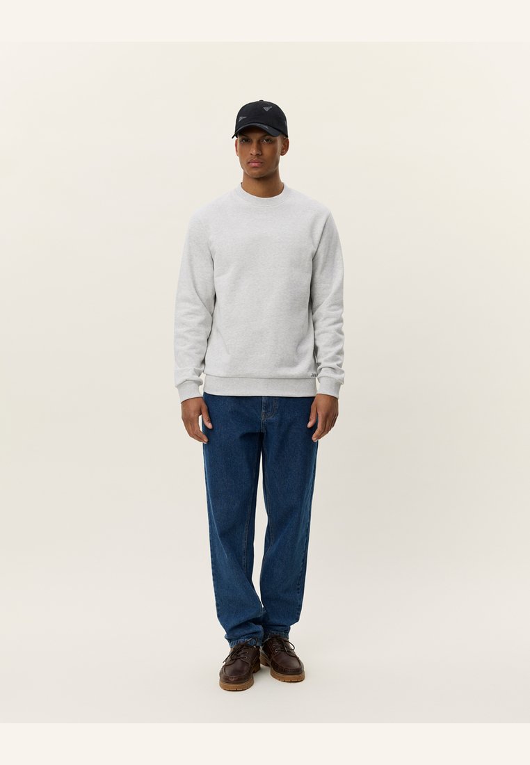 Sudadera gris con cuello redondo, de tela ligera y puños de punto, combinada con unos vaqueros de denim azul holgados y zapatos marrón oscuro. Gorra negra con logo.