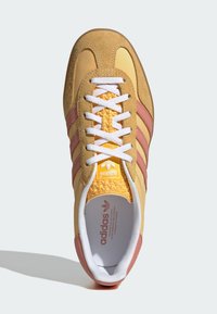 Zapatilla amarilla y beige con franjas rosas, parte superior de ante y sintético, cordones blancos, lengüeta texturizada y logotipo en el talón y la lengüeta.