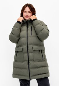 Manteau matelassé vert olive avec fermeture éclair et bouton-pression, comprenant deux poches avant et une poche poitrine zippée, en tissu texturé, avec une capuche.