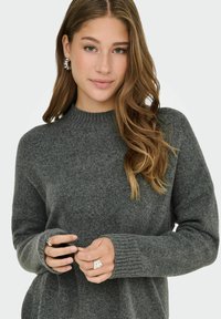 Pull en tricot gris avec un col rond côtelé, des manches longues et une coupe décontractée. Le modèle porte des bagues et des boucles d'oreilles en argent, mettant en valeur des détails texturés.