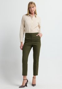 Beige Button-up-Hemd mit einer taillierten Silhouette, kombiniert mit olivgrünen, hochgeschnittenen Hosen und braunen Absatzschuhen.