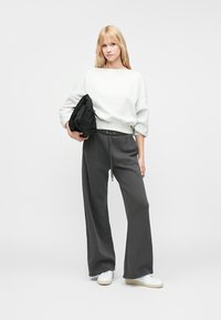 Sivi oversized dukserica sa širokim rukavima, uparena s tamnosivim širokim hlačama i bijelim tenisicama. Model drži crnu torbu.