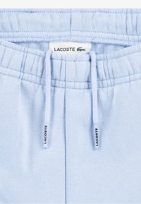 Svetlomodré tepláky s elastickým pásom, sťahovacími šnúrkami a štítkom Lacoste. Mäkká textúra a viditeľné švy.