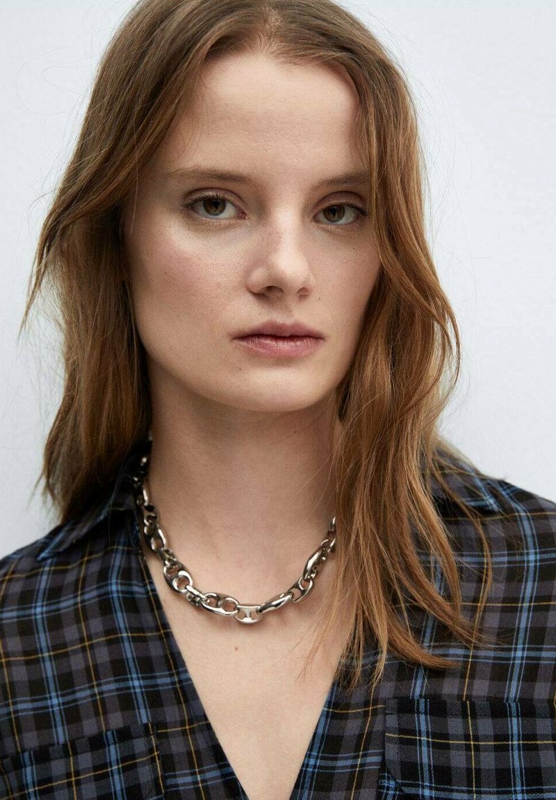 Camicia a quadrati neri e blu con colletto, decorata con una collana di catena metallica spessa. I capelli sono ondulati e di lunghezza fino alle spalle. Sfondo semplice.
