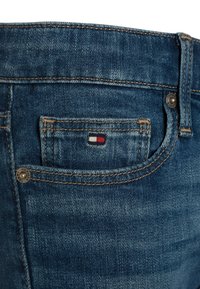 Denim farkut tummansinisen pesun kanssa, joissa on takatasku, jossa on pieni brodeerattu logo punaisella, valkoisella ja sinisellä, sekä näkyvätomminkaivopistokkaat.