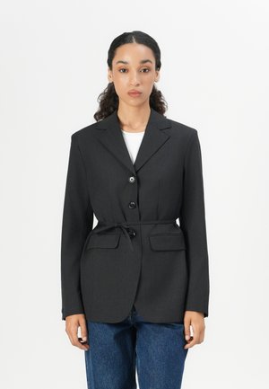 Black tailored blazer with a fitted waist, notched lapels, a two-button front, and two front pockets. Worn over a white shirt.