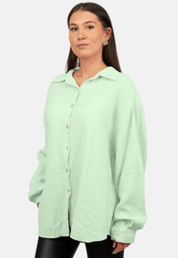 Camicia button-up verde chiaro in tessuto texturizzato con colletto, maniche lunghe e vestibilità ampia, abbinata a leggings neri.
