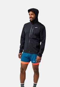 Črna jakna z zadrgo in kapuco, ki ima žep na prsih in elastične manšete, v kombinaciji z modrimi kratkimi hlačami in vzorčanimi nogavicami.