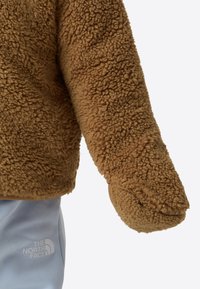 Mâneca jachetei maro din fleece pufos și pantaloni albastru deschis cu logo-ul alb The North Face pe fundal simplu.