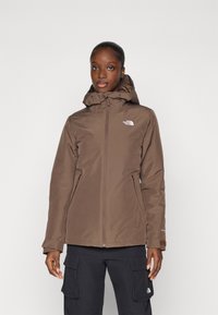 Bruine geïsoleerde jas met een ritssluiting en een verstelbare kap. Voorzien van het North Face-logo op de borst en twee zijzakken.
