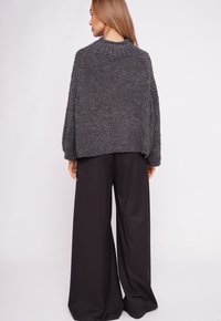 Kobieta o długich włosach, ubrana w ciemnoszary sweter o fakturze oraz szerokie czarne spodnie, stojąca tyłem na jednolitym tle.