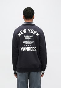 Tengerészkék baseball dzseki, amelyen a hátoldalon fehérrel a „NEW YORK YANKEES” felirat szerepel, csíkos gallérral és mandzsettával, valamint puha tapintású anyagból.