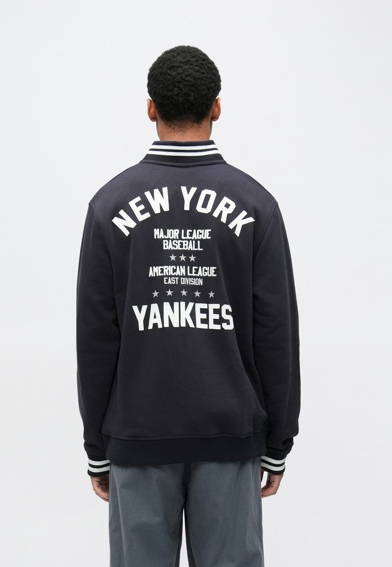 Tengerészkék baseball dzseki, amelyen a hátoldalon fehérrel a „NEW YORK YANKEES” felirat szerepel, csíkos gallérral és mandzsettával, valamint puha tapintású anyagból.