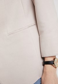 Veste côtelée rose clair avec une poche et une silhouette élégante. Une montre noire à cadran rond complète la tenue.
