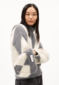 Gestrickter Pullover mit hohem Kragen, der ein geometrisches Muster in Grau und Creme zeigt, lockere Ärmel und eine strukturierte Oberfläche aufweist. Etikett am Ärmel.