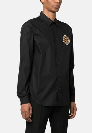 Camicia nera con bottoni in tessuto leggero a maniche lunghe, dotata di una toppa rotonda con logo dorato sul lato sinistro del petto. Finitura liscia e strutturata.