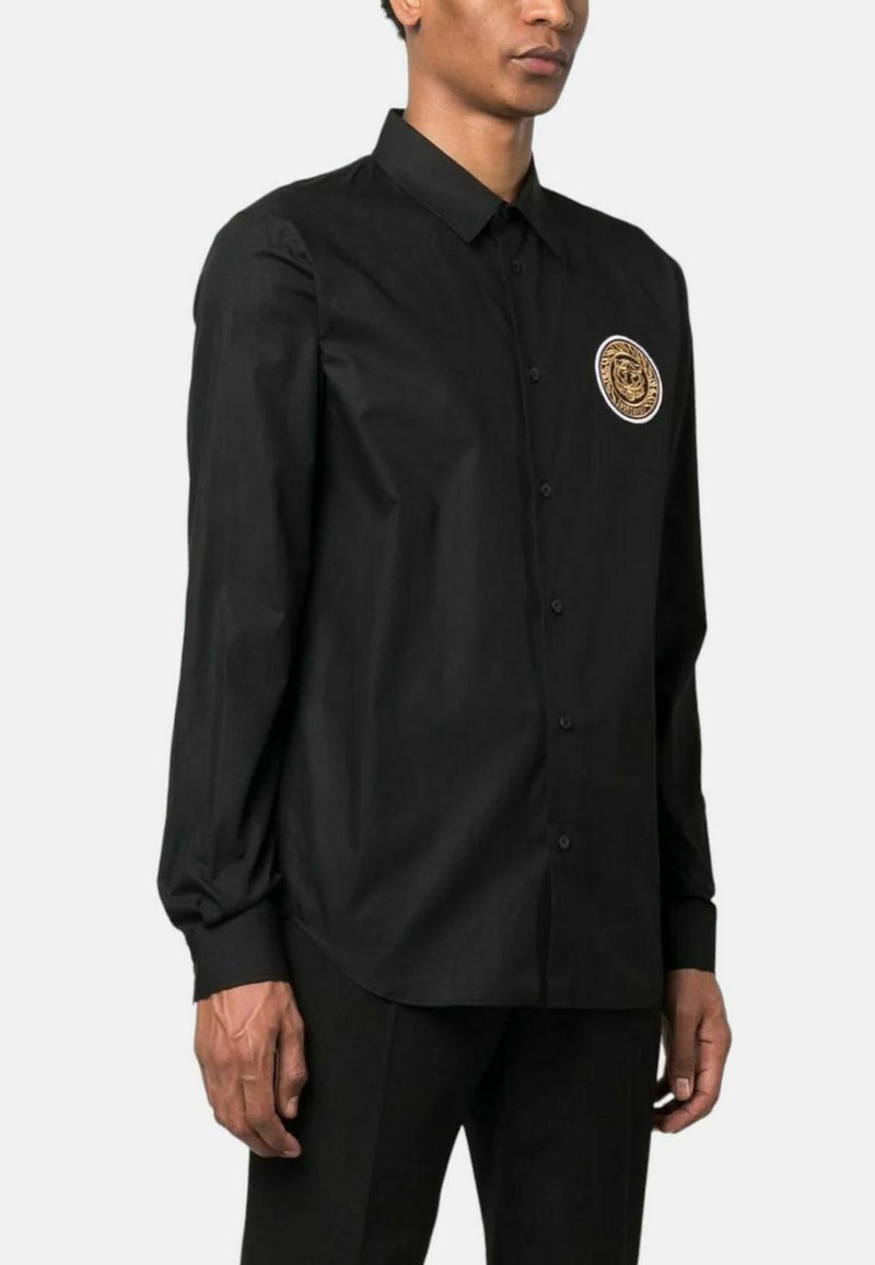 Camicia nera con bottoni in tessuto leggero a maniche lunghe, dotata di una toppa rotonda con logo dorato sul lato sinistro del petto. Finitura liscia e strutturata.