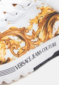 Λευκό αθλητικό παπούτσι με χρυσό σπιράλ σχέδιο και μαύρη σόλα. Διαθέτει υφή στην επάνω επιφάνεια και εμφανές λογότυπο "VERSACE JEANS COUTURE."