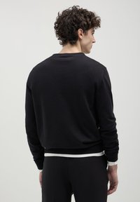 Uomo con capelli ricci che indossa un maglione nero a maniche lunghe sopra una camicia bianca, visto da dietro contro uno sfondo chiaro e semplice.
