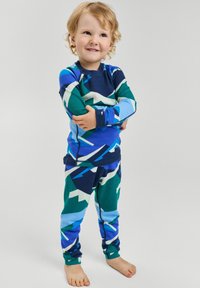 Piżama z długim rękawem w niebieskie, zielone i białe geometryczne wzory; miękki materiał o dopasowanym kroju. Zestaw zawiera pasujące spodnie. Dziecko stoi ze skrzyżowanymi ramionami.
