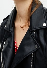 Veste de moto en simili cuir noir avec des boutons-pression argentés, une fermeture éclair diagonale et un large col. Superposée sur une chemise à fleurs rouges.