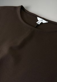 Hnedá polovičná košeľa vyrobená z hladkej tkaniny, so zaobleným výstrihom a s etiketou s názvom značky a veľkosťou umiestnenou na krku.