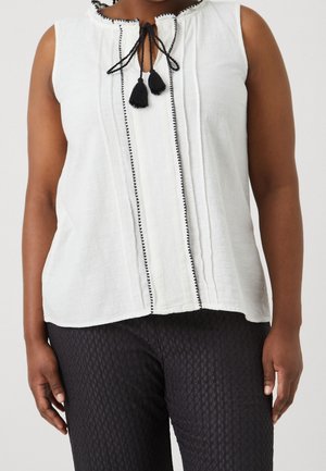 Witte mouwloze top met zwarte afwerking, gekruiste kraag en keyhole sluiting met kwastjes. Geweven stof met verticale plooien. Gecombineerd met gedessineerde donkere broek.