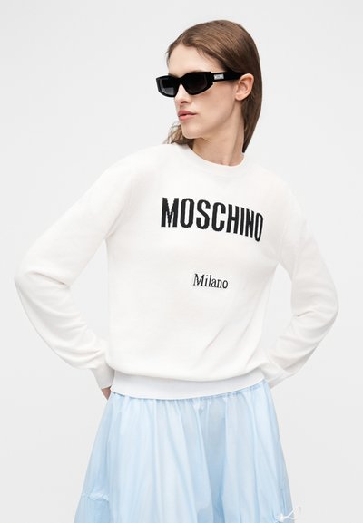 Γυναίκα που φοράει μαύρα γυαλιά ηλίου Moschino, λευκό πουλόβερ με το κείμενο "MOSCHINO Milano" και ανοιχτόχρωμη μπλε φούστα, ποζάροντας με τα χέρια στη μέση.