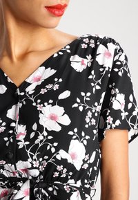 Abito nero floreale con grandi fiori bianchi e cuori rosa. Tessuto morbido, maniche corte, scollo a V e dettaglio con cintura legata in vita.
